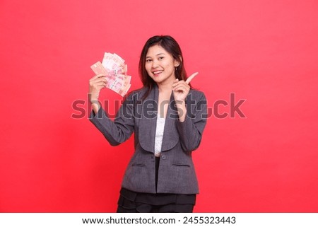 Asian office girl smiling expression holding credit, debit card and money while pointing top left at the camera wearing gray jacket and red skirt. for transaction, business and advertising concepts