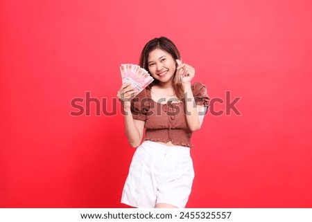 Cheerful indonesia woman gestures as a sign of love (saranghaeyo) while holding rupiah currency wearing a brown blouse with a red background. for transaction, technology and advertising concepts