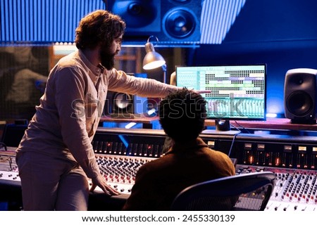 Skilled musician working with audio engineer to edit new recorded tracks in control room, pressing buttons and faders on panel board desk. Team of artist and producer creating music in studio.