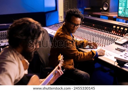 Artist playing a new record on his guitar in control room, recording and producing music with audio engineer in studio. Musician singing on electro acoustic instrument next to mixing console.