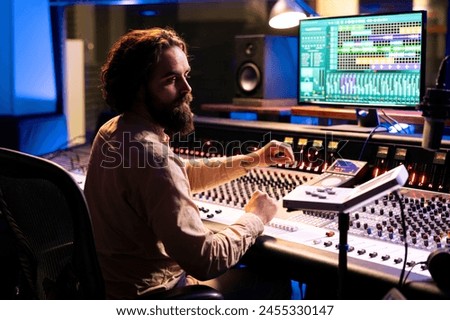 Music technician mixing and mastering songs with motorized faders and knobs, using control desk dashboard and computer. Young sound engineer creating tracks for an album in studio.