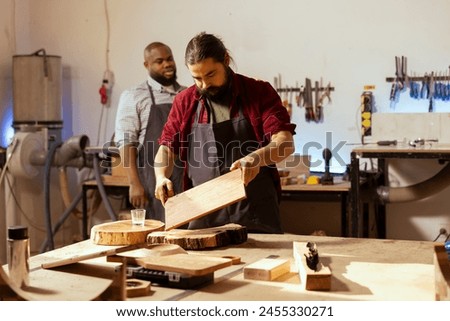 Craftsperson at workbench starting restoration project on wood block next to african american coworker. Carpenter preparing to color plank with paintbrush near apprentice solving tasks behind Royalty-Free Stock Photo #2455330271