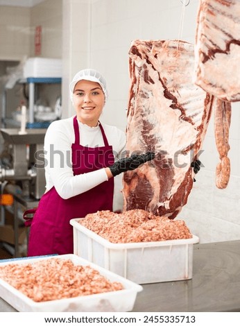 Positive young butcher shop saleswoman offering fresh raw beef ribs at meat processing table..