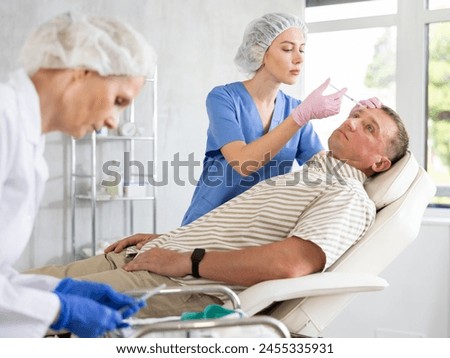 Team of cosmetologists provide anti-aging injection services to male patient. Young female doctor performs wrinkle smoothing procedure, gives injection of botulinum toxin to man patient in forehead Royalty-Free Stock Photo #2455335931