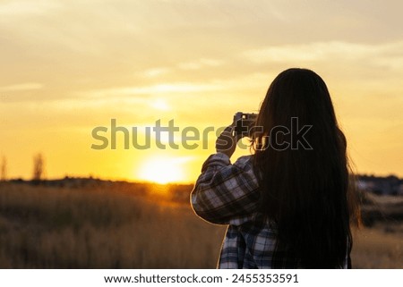 Young woman in checkered shirt taking photos with an analog camera in the field at sunset. The sky is orange and the colors in the image convey tranquility.