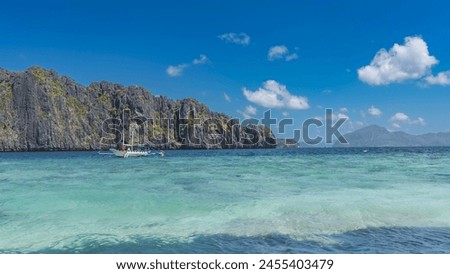 The traditional Filipino double-outrigger dugout bangka boat is anchored in the turquoise ocean. Picturesque steep karst cliffs against a blue sky and clouds. An island in the distance. Philippines. 