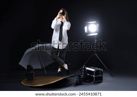 Female photographer with modern camera and lighting equipment taking picture of viewer on black background