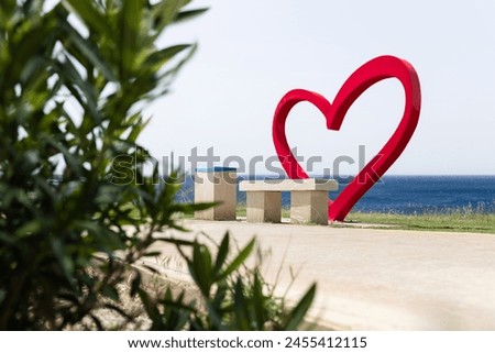 A red heart stands on the coastline of the Mediterranean Sea in Paphos. Next to it is a bench. The heart, made of metal, symbolizes love and family, serving as a spot for wedding photoshoots. Royalty-Free Stock Photo #2455412115