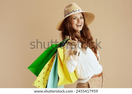 Beach vacation. smiling stylish middle aged woman in white blouse and shorts isolated on beige background with shopping bag and straw hat. Royalty-Free Stock Photo #2455417959