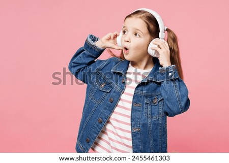 Little child shocked cute kid girl 7-8 years old wears denim shirt listen to music in headphones look aside on area isolated on plain pastel pink background. Mother's Day love family lifestyle concept