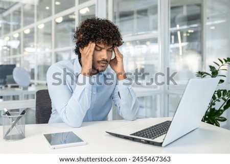 A young man experiences stress while working on his laptop at a modern, well-lit office. He is displaying signs of headache or concentration difficulty.