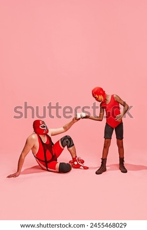 Masked wrestler in red gear seated, toasting milkshakes with standing partner against pink studio background. Concept of pop art, generation difference, costume festivals, competitions. Ad