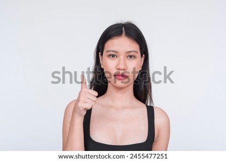 Young Asian woman in a black bodysuit showing approval with a thumbs up gesture, isolated on a white background.