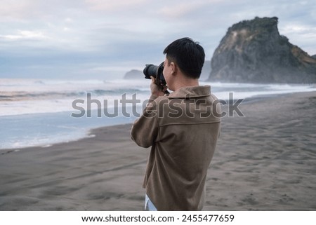 A casually dressed male tourist is standing and taking a photo with iconic Lion Rock on Piha Beach in the evening at West of Auckland, New Zealand