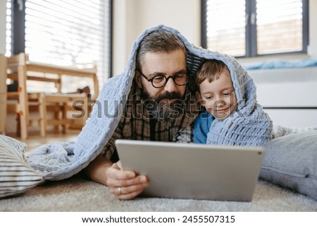 Little boy watching cartoon movie on tablet with father, lying under blanket on floor in kids room. Dad explaining technology to son, digital literacy for kids. Royalty-Free Stock Photo #2455507315