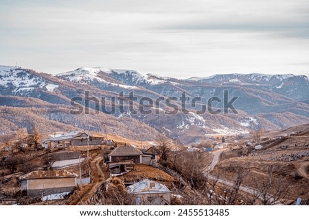 picturesque natural picture with snow-capped huge mountain peaks and mountains, autumn bare trees, gray sky, snow lies on the tops of the mountains and abandoned old destroyed houses in the village of