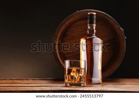 Whiskey with ice cubes in glass, bottle and barrel on wooden table against black background, space for text