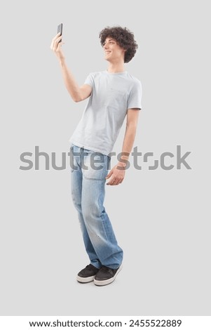 Young happy smiling handsome man using his smartphone, taking a selfie isolated against a gray background, full body image