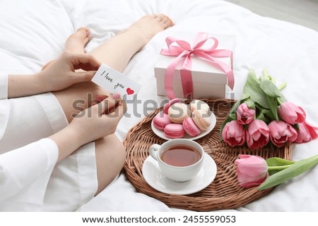 Tasty breakfast served in bed. Woman with I Love You card, macarons, tea, flowers and gift box at home, closeup