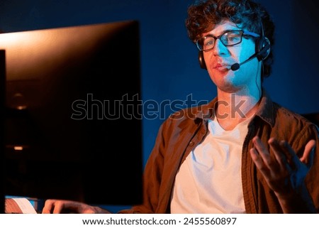 Young creative businessman communicating with listeners on laptop screen or meeting with team member, wearing headphones at neon light modern home office. Concept of working at night time. Gusher.
