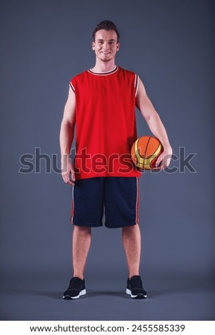 Full-length image of handsome young basketball player in uniform holding a ball, looking at camera and smiling, on gray background Royalty-Free Stock Photo #2455585339