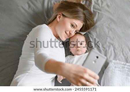 Mother lying on bed with her newborn takes photo, takes selfie using a smartphone, top view.Mom calls using video call to father or relative. Concept of technology, family, memories.