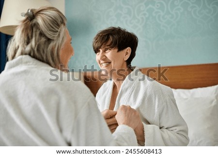 A woman in a bathrobe smiles as another woman puts on her robe in a tender moment of connection. Royalty-Free Stock Photo #2455608413