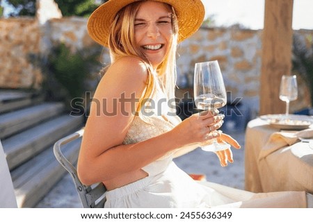 Young woman in a straw hat holds a glass of champagne and smiles sweetly. A blonde in a white dressy sundress drinks sparkling wine in an open-air cafe. An attractive laughing girl in a restaurant. Royalty-Free Stock Photo #2455636349