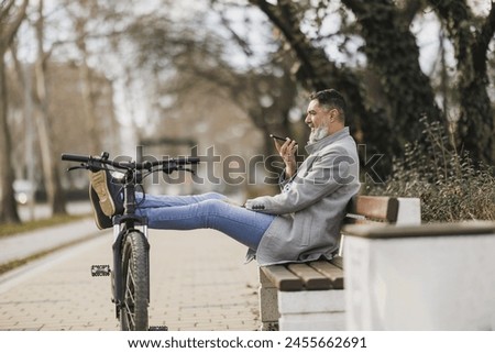A middle-aged man with a beard is sends voice message while taking a break on a bench, with his bicycle propped beside him. Royalty-Free Stock Photo #2455662691