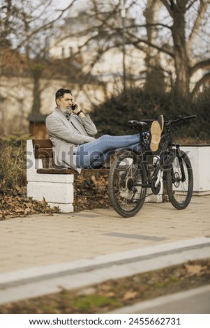 A casual business man takes a break on a bench in a park with his bicycle propped beside him. He is talking on his smartphone. Royalty-Free Stock Photo #2455662731