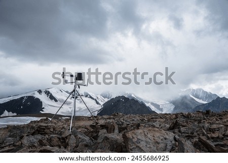 Automatic meteorological station on stone hill with view to large snow-capped mountain range. Measurement speed and direction of wind among glaciers. Measuring atmospheric pressure and precipitation. Royalty-Free Stock Photo #2455686925