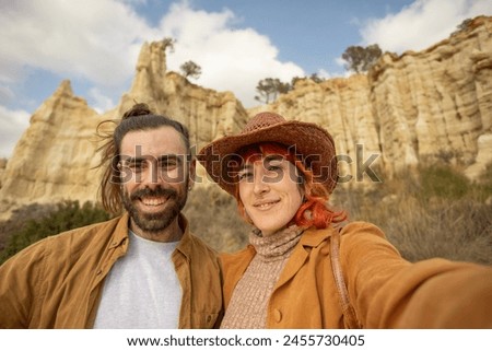 A man and a woman are posing for a picture in front of a mountain. The man is wearing a brown jacket and the woman is wearing a brown hat. Scene is cheerful and lighthearted, as the couple is smiling