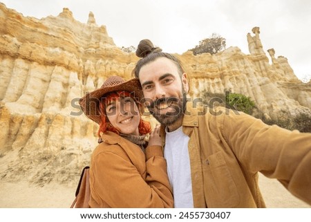 A man and a woman are posing for a picture in front of a mountain. The man is wearing a brown jacket and the woman is wearing a brown hat. They both have smiles on their faces