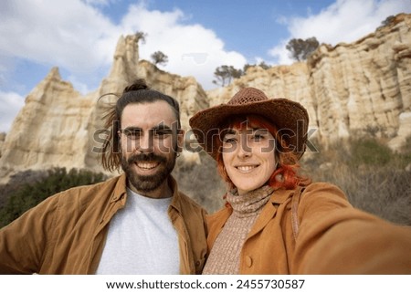 A man and a woman are posing for a picture in front of a mountain. The man is wearing a brown jacket and the woman is wearing a brown hat. They both have a smile on their faces