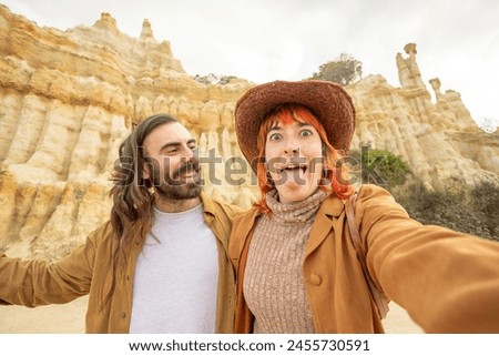A man and a woman are posing for a picture in front of a large rock formation. The man is wearing a brown jacket and the woman is wearing a brown hat. They both have a smile on their faces
