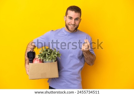 Young caucasian making a move while picking up a box full of things isolated on yellow background making money gesture