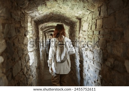 Curious tourist woman standing with backpack in stone passage of ancient castle. View from the back