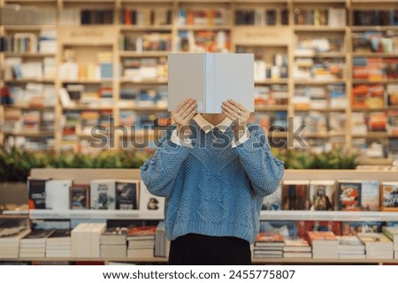 A young woman holds a book in front of her face cover, standing amidst a vibrant and varied bookstore. Shelves of books create a cozy, intellectual backdrop. Royalty-Free Stock Photo #2455775807