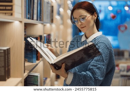 A focused young woman with glasses reads a large book amidst shelves filled with books in a cozy, well-lit bookstore. The scene exudes a calm and intellectual atmosphere.