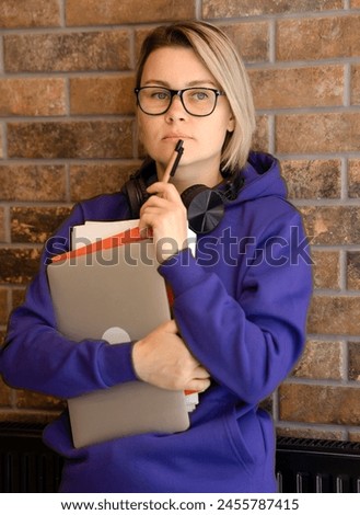 A blonde woman in glasses holds a laptop and papers with her hands, a pen near her face. Girl thinking, brick wall, student with books and textbooks, headphones around her neck, purple sweater