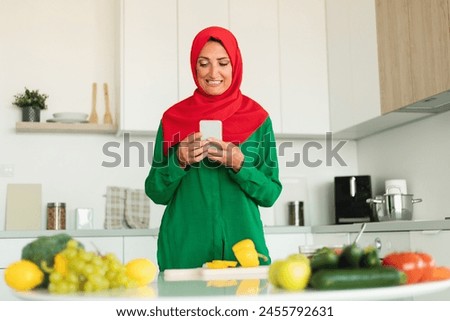 Muslim woman is standing in a modern kitchen, attentively looking at her cell phone screen. The kitchen counter is visible with various appliances and utensils neatly arranged