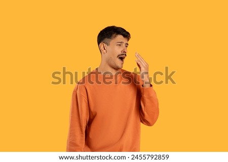 A young man with dark hair stands against a vivid yellow backdrop, covering his mouth with his hand as he yawns widely, displaying signs of fatigue or boredom wearing a casual orange sweatshirt.