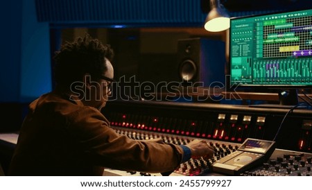 Sound designer uses mixing console and managing audio tracks, twisting knobs on control desk for adjusting tunes and volume. Producer creates music in professional studio, technical gear. Camera B.