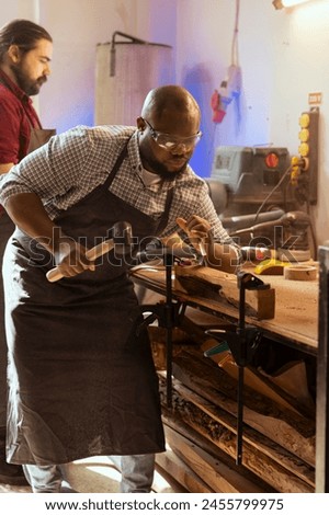Craftsperson carving designs into wood using chisel and hammer next to coworker. Artisan shaping wooden pieces using protective equipment to prevent workplace accidents, helped by apprentice Royalty-Free Stock Photo #2455799975