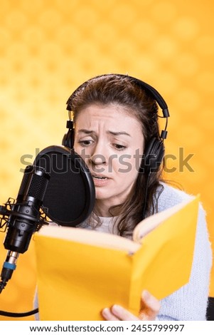 Woman with frown on face wearing headphones reading aloud from book into mic against background. Voice actor recording audiobook, creating engaging content for listeners, glowering for dramatic effect