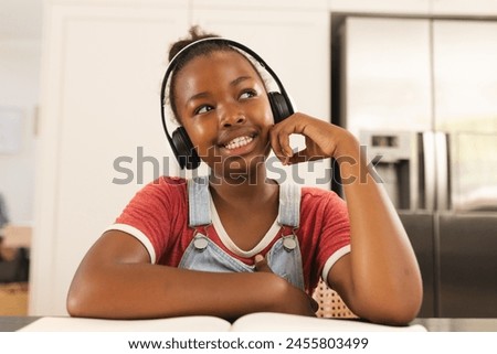 An African American girl wearing headphones, smiling during video call at home. Wooden frames add warmth to a cozy room with a brown background, creating a peaceful atmosphere, unaltered Royalty-Free Stock Photo #2455803499