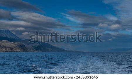 The foam trail behind the stern of the ship stretched across the surface of the blue endless ocean. Ripples on the water. Mountains against the sky and clouds. Beagle Channel.  Argentina.