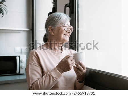 Happy middle aged senior retired woman standing near window, looking in distance, recollecting good memories enjoying peaceful moment drinking a coffee cup Royalty-Free Stock Photo #2455913731