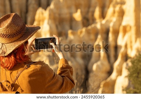 A woman wearing a straw hat is taking a picture of a mountain. The photo is in color and the woman is holding a cell phone