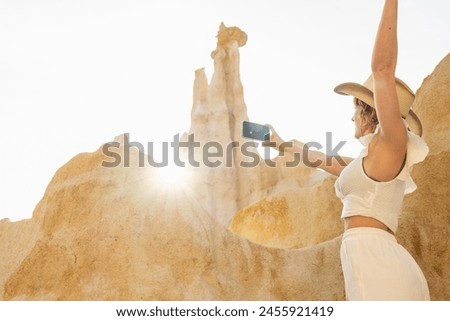 A woman is taking a picture of a mountain with a cell phone. She is wearing a cowboy hat and white shirt